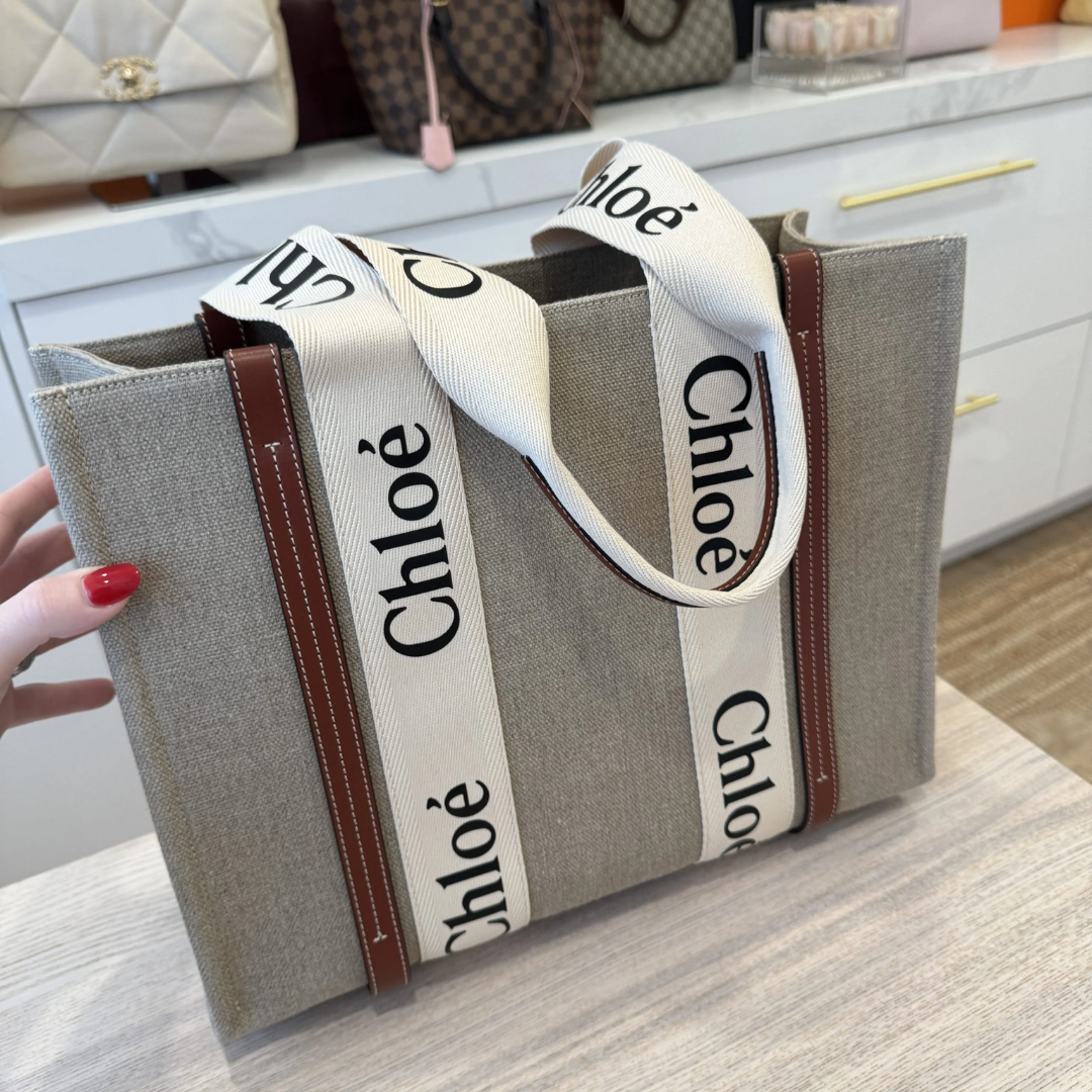 Grey canvas tote bag with white logo straps and brown accents, displayed in a boutique setting.
